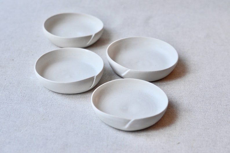 May include: Four white ceramic bowls with a unique, slightly uneven rim. The bowls are arranged in a staggered pattern on a light gray fabric surface.