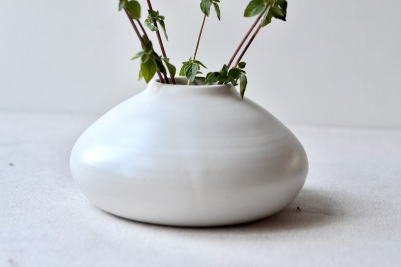 May include: A white ceramic vase with a rounded, organic shape. The vase is filled with green leafy stems.