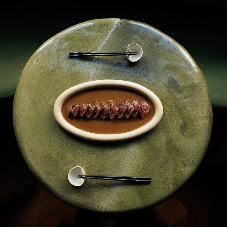 May include: A close-up of a ceramic dish with a brown sauce and thin slices of meat. The dish is on a green table with two small white bowls and two black chopsticks.