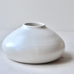 May include: A small, white ceramic vase with a rounded, bulbous shape. The vase has a smooth, glossy finish.