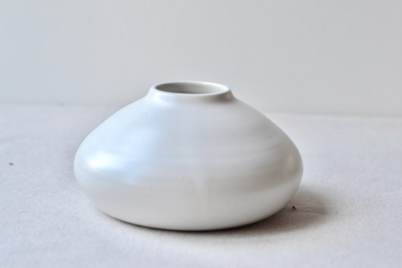 May include: A small, white ceramic vase with a rounded, bulbous shape. The vase has a smooth, glossy finish.