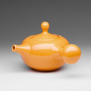 May include: A bright orange ceramic teapot with a rounded body and a bulbous spout. The teapot has a smooth, glossy finish.