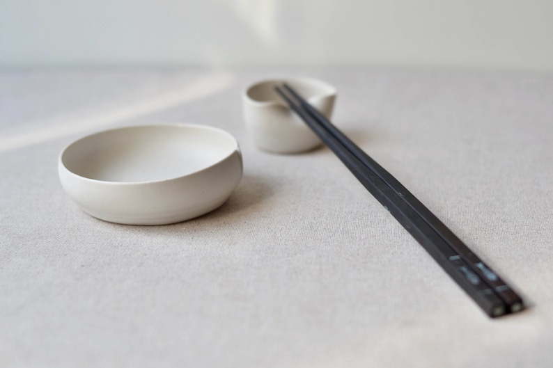 May include: A white ceramic bowl and a small white ceramic dish with a pair of black chopsticks resting on the dish.