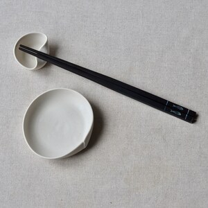 May include: A white ceramic chopstick rest shaped like a leaf, and a pair of black chopsticks with a fish design on them.