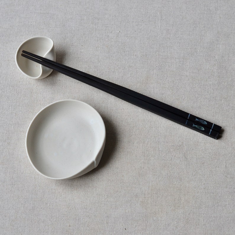 May include: A white ceramic chopstick rest shaped like a leaf, and a pair of black chopsticks with a fish design on them.
