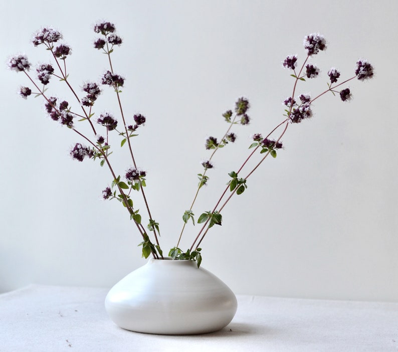 May include: A white ceramic vase with a rounded, organic shape holds a bouquet of purple and white flowers with green stems.