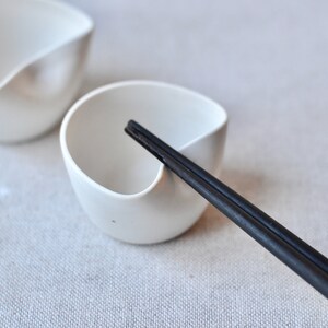 May include: Two white ceramic bowls with a unique curved design, one with a pair of black chopsticks resting inside.