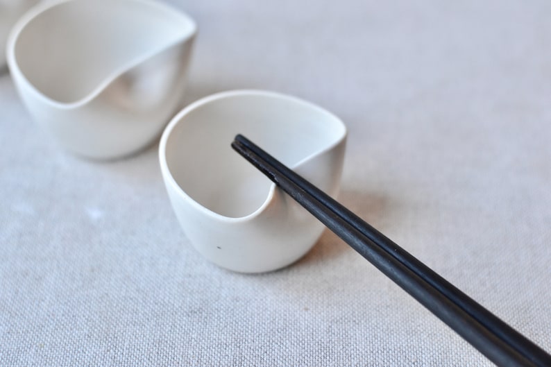May include: Two white ceramic bowls with a unique curved design, one with a pair of black chopsticks resting inside.