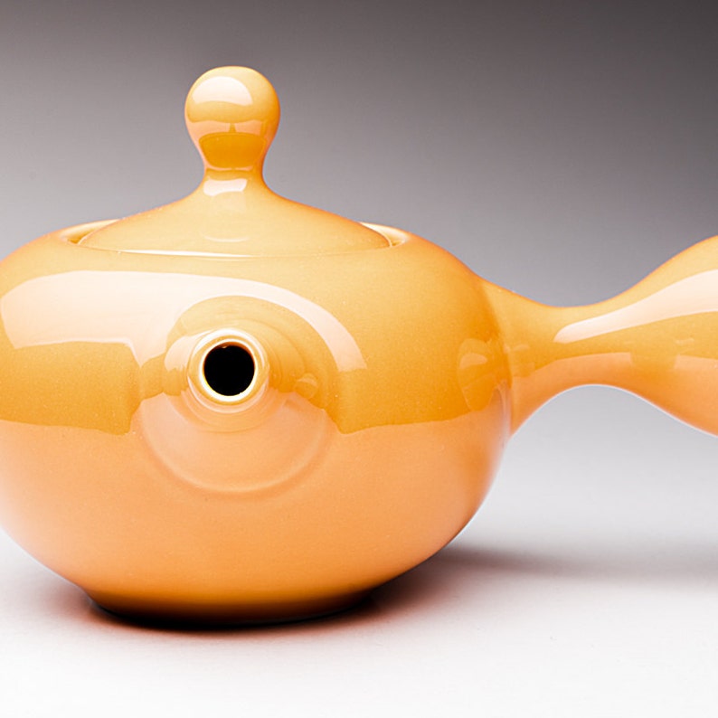 May include: A bright orange ceramic teapot shaped like a fish with a rounded lid and a spout on the side.