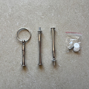 Könnte beinhalten: Silberfarbenes Metall-Kuchenständer-Hardware-Set. Enthält einen Griff, zwei zentrale Stützstangen und einen kleinen Beutel mit Schrauben und Unterlegscheiben. Die Teile sind für den Zusammenbau eines mehrstöckigen Servierständer konzipiert.