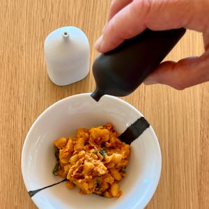 Puede incluir: Un cuenco de cerámica blanco y negro lleno de un plato colorido. Una botella negra vierte líquido sobre la comida. Un recipiente de cerámica blanco se encuentra sobre una superficie de madera.