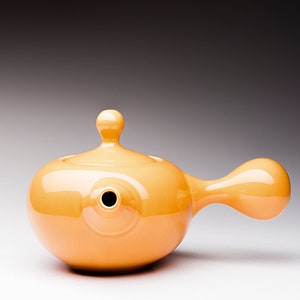 May include: A bright orange ceramic teapot with a rounded body and a long, curved spout. The teapot has a small, rounded lid with a knob on top.