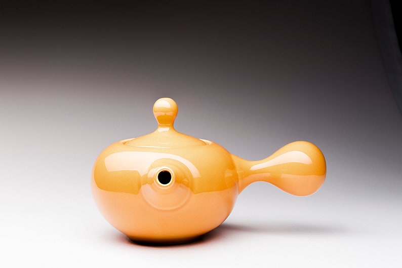 May include: A bright orange ceramic teapot with a rounded body and a long, curved spout. The teapot has a small, rounded lid with a knob on top.