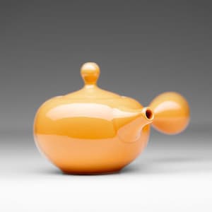 May include: A bright orange ceramic teapot with a rounded body and a spout on the side. The teapot has a unique design with a rounded handle.