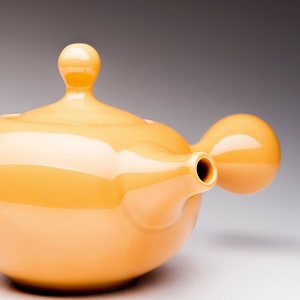 May include: A yellow ceramic teapot with a rounded body and a spout on the side. The teapot has a lid with a rounded knob on top.