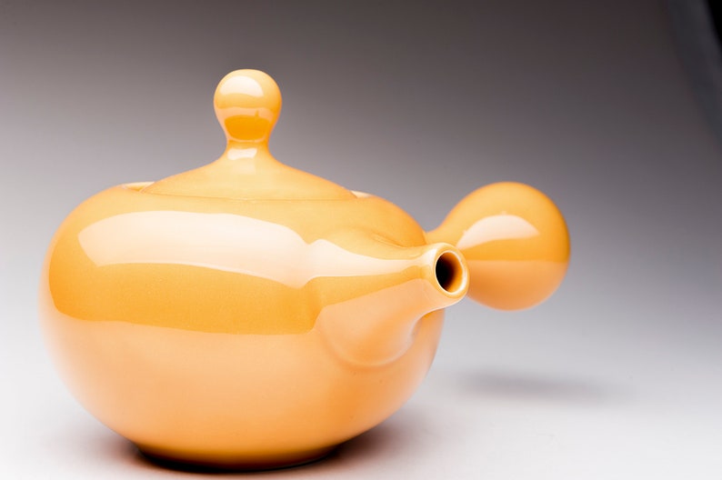 May include: A yellow ceramic teapot with a rounded body and a spout on the side. The teapot has a lid with a rounded knob on top.