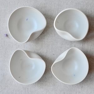May include: Four white ceramic bowls with a unique, slightly asymmetrical shape. The bowls are smooth and have a subtle, off-white color.