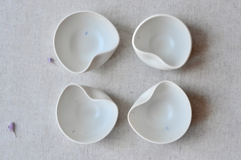May include: Four white ceramic bowls with a unique, slightly asymmetrical shape. The bowls are smooth and have a subtle, off-white color.