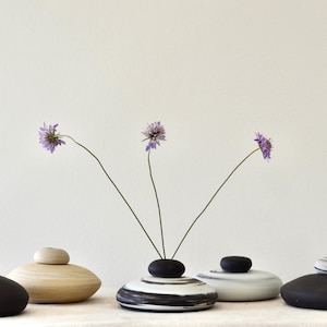 May include: A collection of five decorative ceramic vases in various colors and designs. The vases are arranged in a row, each with a unique shape and a small, rounded lid. Three purple flowers are placed in the center vase.