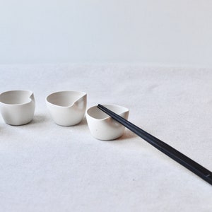 May include: Four white ceramic bowls with a unique, asymmetrical design, arranged in a row on a white surface. A pair of black chopsticks are placed diagonally across the surface, resting on one of the bowls.