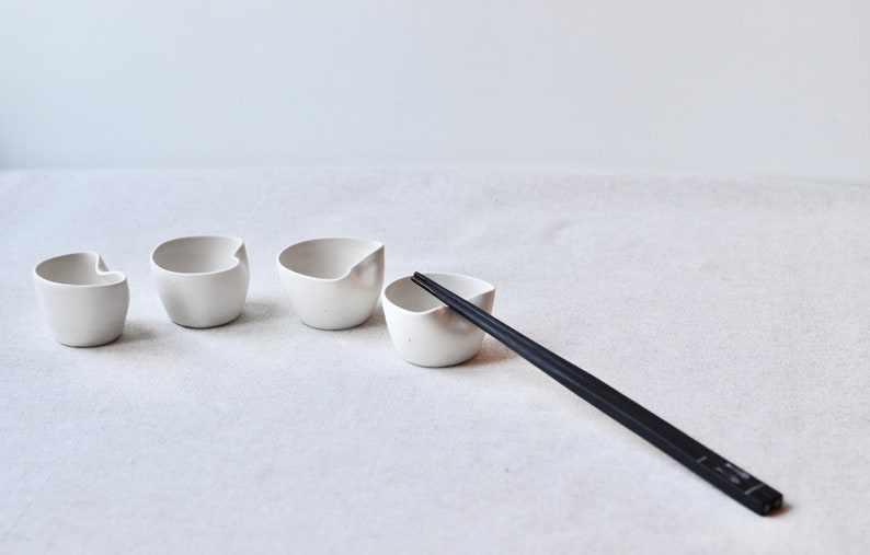 May include: Four white ceramic bowls with a unique, asymmetrical design, arranged in a row on a white surface. A pair of black chopsticks are placed diagonally across the surface, resting on one of the bowls.