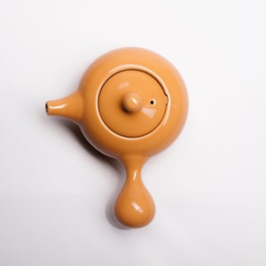 May include: A small, orange ceramic teapot with a rounded body and a single spout. The teapot has a lid with a small knob on top.
