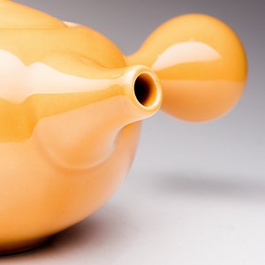 May include: A yellow ceramic teapot with a rounded body and a spout. The teapot has a unique design with a bulbous shape on the side.