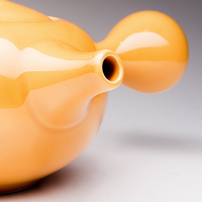 May include: A yellow ceramic teapot with a rounded body and a spout. The teapot has a unique design with a bulbous shape on the side.