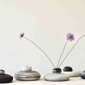 Puede incluir: Una colección de pequeños jarrones decorativos en tonos negro, gris y blanco. Cada jarrón tiene una base redondeada en forma de piedra y una pequeña parte superior contrastante. Delicadas flores moradas con finos tallos están dispuestas en algunos jarrones.