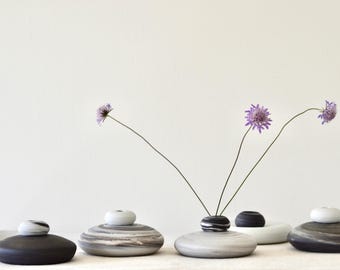 May include: A collection of small, decorative ceramic vases in shades of black, gray, and white. Each vase has a rounded, stone-like base and a small, contrasting top. Delicate purple flowers with thin stems are arranged in some of the vases.