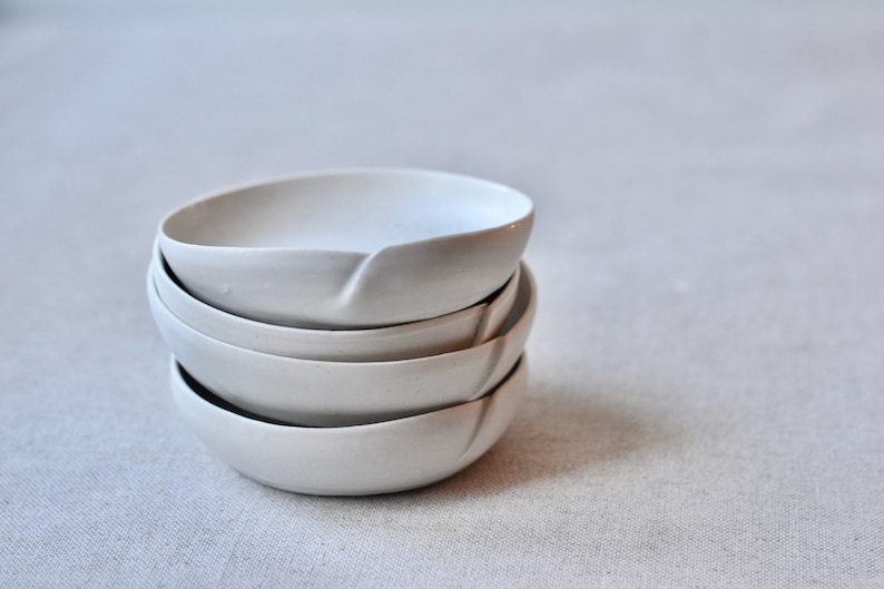May include: A stack of four white ceramic bowls with a unique, slightly curved rim. The bowls are nested together, creating a visually appealing display.