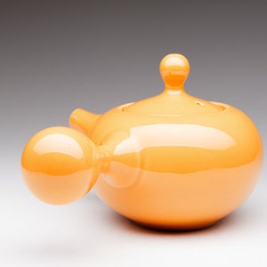 May include: A bright orange ceramic teapot with a rounded body and a bulbous spout. The teapot has a smooth, glossy finish.