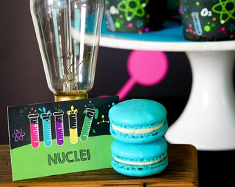 May include: A science-themed party table setting with a light bulb, cupcakes decorated with atom symbols, and blue macarons. The table has a sign that says "NUCLEI" with test tubes filled with colourful liquids.