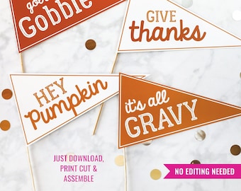 May include: Four pennant-shaped signs with wooden sticks. The signs are orange and white with phrases like "Gobble," "Give thanks," "Hey Pumpkin," and "It's all Gravy." Gold confetti dots the background. Text reads "Download & Print Today!" and "Just Download, Print Cut & Assemble."