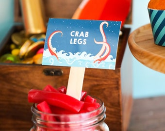 May include: A jar of red licorice sticks with a sign reading "Crab Legs". A wooden treasure chest filled with gold coins and a wooden tray with cupcakes are in the background. The scene is set on a wooden table.