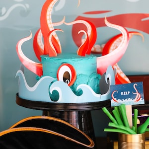 May include: A blue and green cake decorated with red and pink paper octopus tentacles. The cake is on a black cake stand and there is a sign that says "KELP" with a small octopus illustration. There are green candy sticks in a gold can.