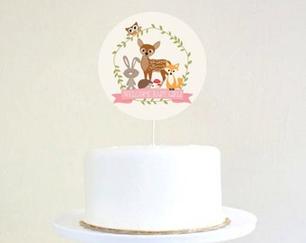 May include: White cake with a round topper featuring a deer, owl, rabbit, fox, and mushrooms. The topper has a pink banner with the text "Welcome Baby Girl".