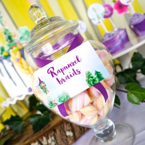 May include: A clear glass candy jar filled with pastel-colored marshmallows. A purple ribbon wraps around the jar, holding a label that reads "Rapunzel braids." The jar sits on a table with other party decorations.