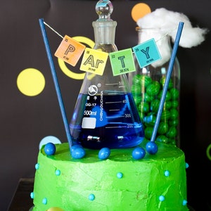 May include: A green and blue frosted cake decorated with blue and green candies. A glass beaker filled with blue liquid sits on top of the cake. A banner with the word "PARTY" spelled out using chemical element symbols hangs above the cake.