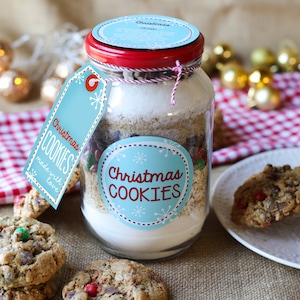 Pode incluir: Um frasco de vidro cheio de ingredientes para biscoitos, rotulado como "Christmas COOKIES". O frasco tem uma tampa vermelha e uma etiqueta azul que diz "Christmas COOKIES made with love". Vários biscoitos assados com doces coloridos estão em frente ao frasco.