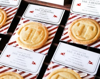 May include: A close-up of several small, round, white cookies with a red and white striped background. The cookies have a design of a ship's wheel on them. The cookies are packaged in clear plastic bags with a white tag that says "Thank you for sail-a-brating with me" and "Spillers & Bakers Pilot Crackers".