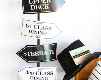 May include: A decorative signpost with black and white directional signs, each pointing left. The signs indicate locations like Ballroom, Upper Deck, and Gymnasium. A black top hat rests on a wooden chair, with a postcard of a ship tucked inside.