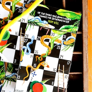Op de afbeelding: Een zwart-wit spelbord met slangen en ladders. Het bord is gemaakt van karton en heeft een glanzende afwerking. De nummers 1 tot en met 31 zijn op het bord gedrukt. Het spel heet "Up the Snake and Down Ladder." De tekst "Thanks for Slithering On By!" en "From Will" is op het bord gedrukt.