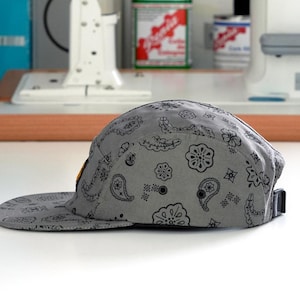 Pu&ograve; includere: Un cappellino grigio a cinque pannelli con un motivo paisley nero. Il cappellino ha una visiera piatta e un cinturino regolabile nero. Il cappellino &egrave; su una superficie bianca.