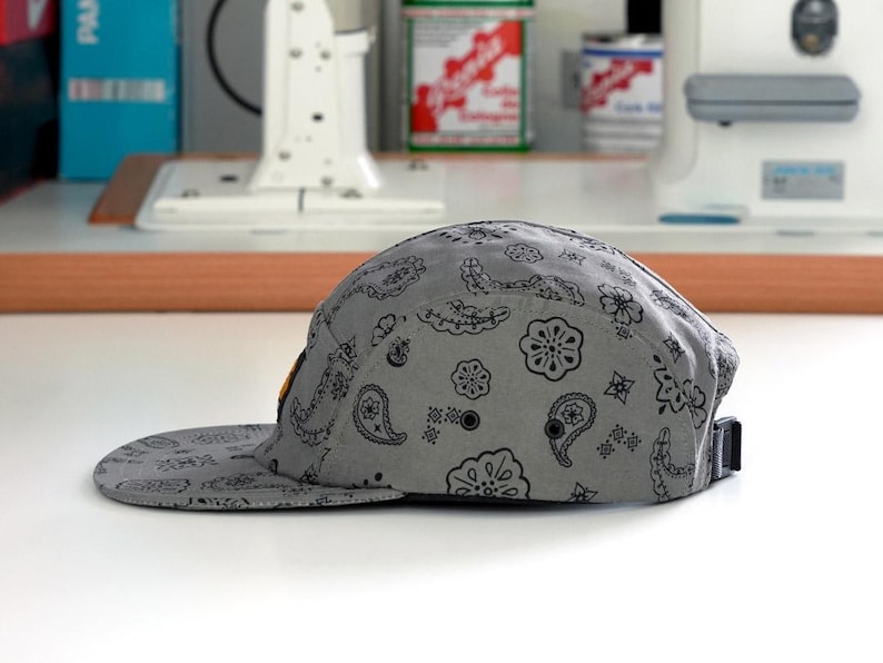 Pu&ograve; includere: Un cappellino grigio a cinque pannelli con un motivo paisley nero. Il cappellino ha una visiera piatta e un cinturino regolabile nero. Il cappellino &egrave; su una superficie bianca.