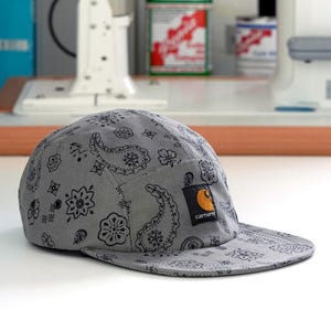 Pu&ograve; includere: Un berretto grigio a cinque pannelli con un motivo paisley nero. Il berretto presenta una piccola toppa quadrata del marchio Carhartt sul davanti. La visiera &egrave; corta e piatta. Il berretto &egrave; su una superficie bianca.