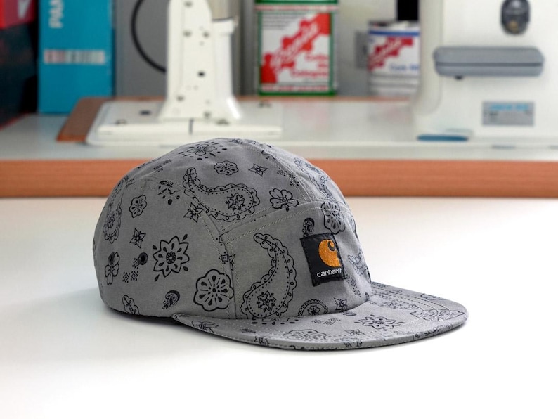 Pu&ograve; includere: Un berretto grigio a cinque pannelli con un motivo paisley nero. Il berretto presenta una piccola toppa quadrata del marchio Carhartt sul davanti. La visiera &egrave; corta e piatta. Il berretto &egrave; su una superficie bianca.