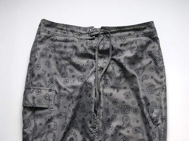 Pu&ograve; includere: Pantaloni cargo grigi con motivo paisley nero. I pantaloni hanno un punto vita con coulisse e una tasca cargo sulla gamba sinistra. Il tessuto ha una leggera lucentezza.