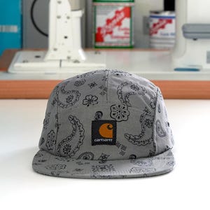 Pu&ograve; includere: Cappellino Carhartt grigio a cinque pannelli con motivo paisley. Il cappellino presenta una toppa quadrata con il logo Carhartt e una 'C' arancione sul davanti. Il cappello &egrave; realizzato in tessuto morbido e ha una visiera piatta.