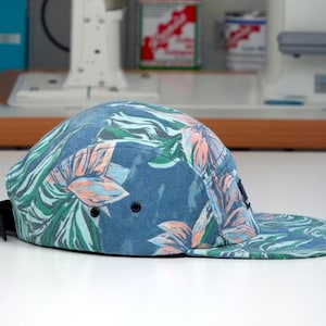 Könnte beinhalten: Blaue 5-Panel-Cap mit flachem Schirm, verziert mit einem tropischen Blumenmuster in Grün, Rosa und Weiß. Sie hat einen schwarzen verstellbaren Riemen und zwei schwarze Ösen. Ein modisches Accessoire.